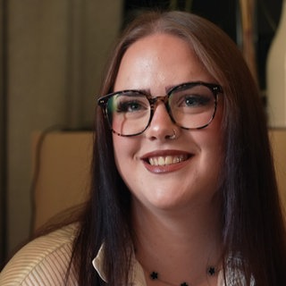 Eine junge Frau mit glatten Haaren und Brille sitzt in einem Raum und lächelt in die Kamera.