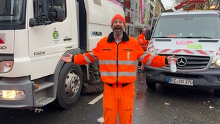 Müllwerker in Arbeitskleidung steht lächelnd mit ausbegreiteten Armen vor Autos.