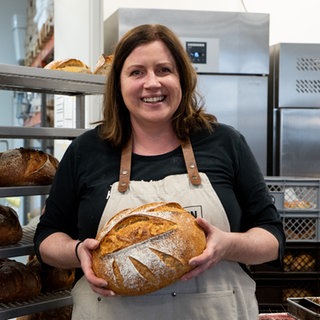 Frau mit braunen Haaren und weißer Schürze steht lächeln in Backstube und hat ein Brot in der Hand.