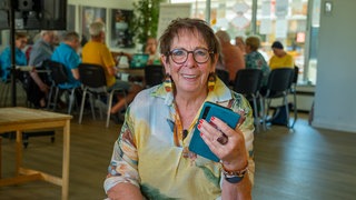 Frau mit braunen Haaren und Brille in buntem Hemd lächelt in die Kamera. In der Hand hat sie ein Handy.