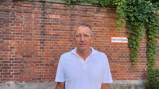 Mann mit grauen Haaren im weißen T-Shirt vor Backsteinmauer.