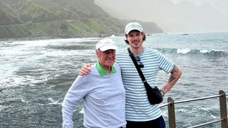 Cedric verbringt viel Zeit mit seinem Opa Gery. Sie befinden sich im Urlaub und im Hintergrund ist ein Gewässer zu sehen. Cedric legt seinen Arm um seinen Opa. Die lachen in die Kamera. 