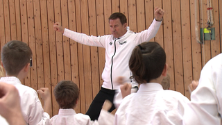 Eugen trägt eine weiße Sportjacke, auf der ‚Trainer‘ steht und macht Kindern in seiner Kampfkunstschule eine Ju-Jitsu-Übung vor.