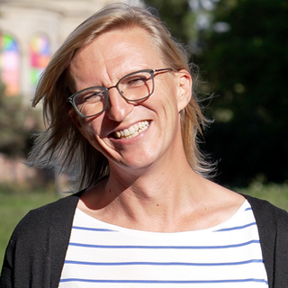Tanja steht in einem Park, hinter ihr eine bunte Villa, sie trägt eine Brille und ein blau-weiß gestreiftes T-Shirt, sie lächelt übers ganze Gesicht. Heute als Frau fühlt sie sich wohl in ihrem Körper. Doch als ihr Vater noch lebte, hat sie sich nicht getraut, sie selbst zu sein. 
