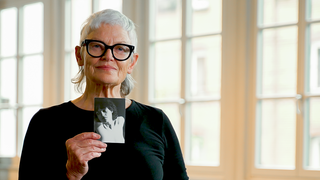 Tänzerin Roswitha (74) steht in einem Tanzstudio und hält ein schwarz-weiß-Foto aus ihrer Jugend in die Kamera, das sie als Teenagerin zeigt.