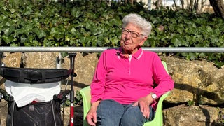 Hildegard sitzt im Garten der Tagespflege in Bad Cannstatt. Die Sonne scheint, sie trägt ein pinkes Polo und eine Brille. Ihr Rollator steht neben ihr, sie sitzt auf einem Gartenstuhl und lächelt.