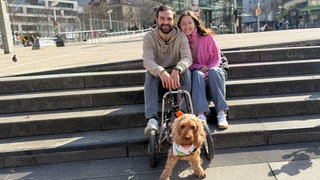 Leonie und Marvin sitzen auf einer Steintreppe und lächeln in die Kamera. Vor den beiden steht Hündin Lilly auf ihren Vorderbeinen. Ihr Rollstuhl hält ihre Hinterbeine.
