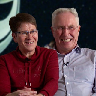 Sigrid und Joachim sitzen nebeneinander und lachen in die Kamera. Im Hintergrund ist verschwommen der Innenraum des Stuttgarter Planetariums zu sehen.