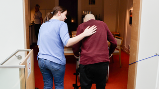 Melanie (links) arbeitet als Pflegerin im Hospiz. Sie läuft mit einem Gast (rechts) zum Esstisch, die beiden sind von hinten zu sehen. Melanies Arm liegt auf dem Rücken des Mannes.