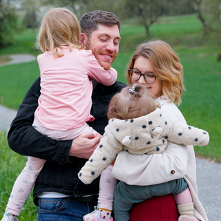 Jenny, ihr Ehemann Sascha und ihre beiden Töchter stehen im Feld. Hinter ihnen ist ein Weg, Bäume und Gras. Sascha hält Ella und Jenny hält Lea im Arm.