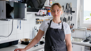 Mariia ist gelernte Köchin und Konditorin. In ihrer Küche steht sie in ihrer Schürze und blickt selbstbewusst in die Kamera. Im Hintergrund sind Schwerlastregale zu sehen, in denen sie ihre Backutensilien aufbewahrt.