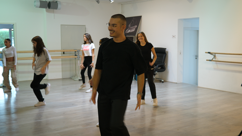 Tanzlehrer Deniz ist in einem Tanzstudio. Dort bringt er Kindern das Tanzen bei. Deniz macht eine Tanzübung vor und lacht dabei. Im Hintergrund sind Kinder zu sehen, die Deniz’ Tanzbewegung nachmachen. 