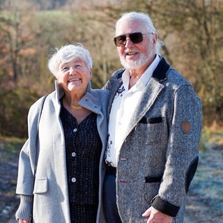 Das Ehepaar Anneliese und Hans stehen im Wald. Sie haben sich in den Armen und lächeln in die Kamera.