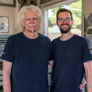 Marcel und sein Papa Rainer stehen im Familienbetrieb. Im Hintergrund sieht man Werkzeug. Bilder von Autos hängen an den Wänden. Marcel hat die Autowerkstatt übernommen. 