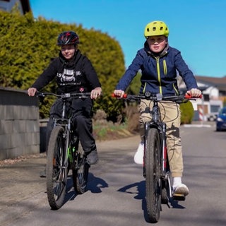 Niklas und Amos fahren jeden Tag mit dem Fahrrad in die Schule. Hier fahren sie gemeinsam durch die Nachbarschaft, hinter ihnen fährt ein Auto.