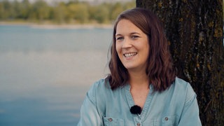 Eine junge Frau mit brauen Haaren sitzt an einen Baum angelehnt, im Hintergrund ist ein See.