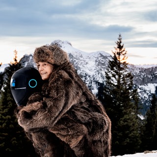 Eine alte Frau im Pelzmantel wird von einem Roboter durch eine schneeverschneite alpine Landschaft getragen (Ausschnitt aus dem Film "Frau Winkler verlässt das Haus") 