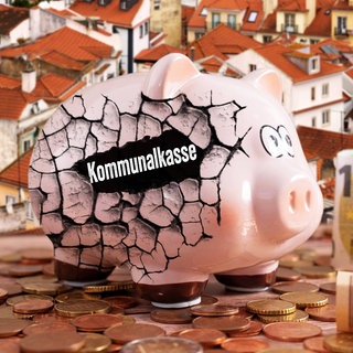 Ein rissiges Sparschwein mit der Aufschrift Kommunalkasse steht auf Münzen vor einem Stadthintergrund symbolisch für klamme Kommunen