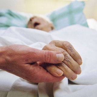 Eine jüngere Person hält die Hand einer alten Person die in einem Bett liegt