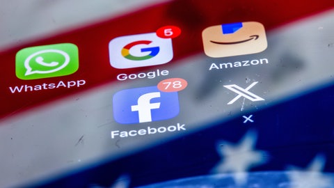 Die Apps verschiedener amerikanischer Tech-Konzerne, WhatsApp, Google, Amazon, Facebook und X, sind auf dem Display eines Smartphones zu sehen, auf dem sich die US-amerikanische Flagge spiegelt.