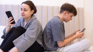 Zwei Jugendliche sitzen Rücken an Rücken auf einem Bett und schauen aufs Handy