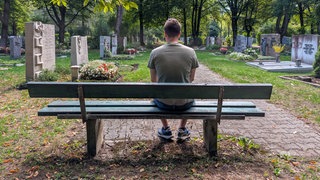 Ein junger Mann sitzt allein auf einer Parkbank auf einem Friedhof und blickt schweigend auf die Grabstätten. 