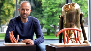 Erwin Wurm sitzt neben seinen Wurst-Skulpturen.