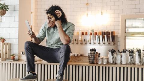 Ein Mann mit Affenmaske schaut auf ein Tablet und sitzt auf einer Theke