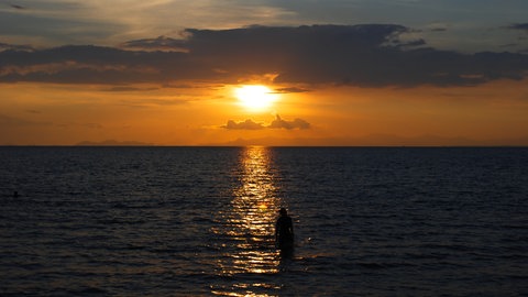 Eine Person steht im Meer während die Sonne untergeht