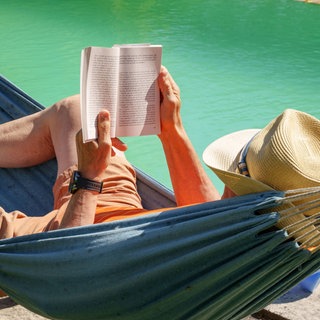 Ein Mann mit einem Sonnenhut liest in einer Hängematte am Pool ein Buch