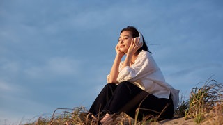 Eine Frau mit Kopfhörer sitzt am Strand auf einer Sanddüne