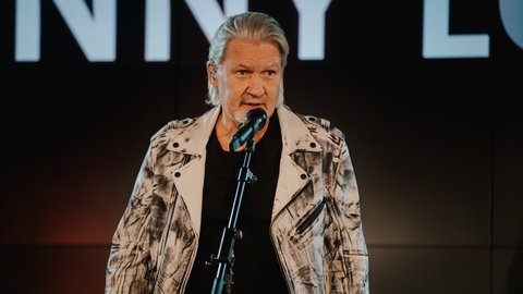 ESC-Legende und Sänger Johnny Logan ("Hold me now") steht beim SWR4 Festival 2020 auf der Bühne und spricht mit dem Publikum. Dabei hat er eine weiß-schwarze Jacke an und ein schwarzes T-Shirt.