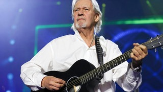 Johnny Logan: Der Sänger und mehrfache Sieger des ESC trägt ein weißes Hemd, passend zu seinem weißen Haar. Er spielt auf einer Gitarre