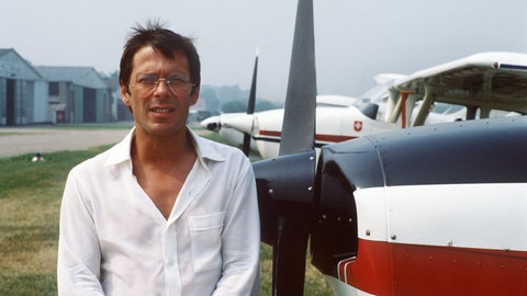 Liedermacher, Chanson-Sänger und Hobbypilot Reinhard Mey ("Über den Wolken") steht in einem kurzärmlichen Hemd neben einem Flugzeug und schaut in die Kamera.