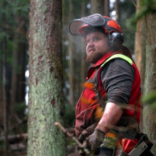 Mario Walz steht im Fränkisch-Schwäbischen Wald und trägt Holzfällerkleidung und einen Schutzhelm.