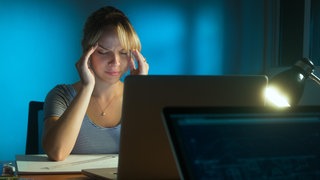 Eine junge Frau sitzt abends gestresst am Schreibtisch in einem Büro | Wie vermeide ich Stress bei einer Urlaubsvertretung?