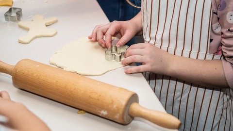 Wellholz neben ausgewellten Salzteig, in dem ein Kind Ausstecher drückt: Zum Herstellen und Trocknen von Deko kann man wie beim Plätzchenbacken vorgehen.
