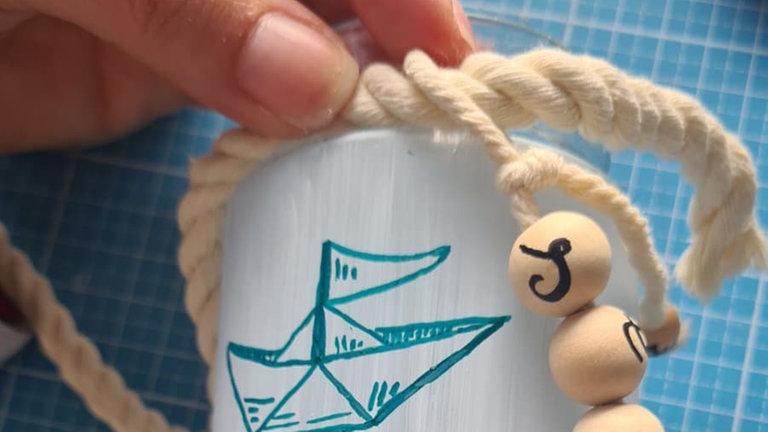 Mit der Schlaufe das Garn an einem hellen Seil befestigen und dieses so zuschneiden, dass es einmal rund um den oberen Glasrand reicht. Anschließend das Seil mit Heißkleber festkleben. 
