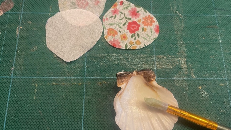 Mit dem Pinsel das innere der Muschel mit Serviettenkleber bestreichen. Das zugeschnittenen Stück Serviette oben passgenau anlegen, mittig von oben nach unten andrücken. Den Rest der Serviette mit dem Pinsel und Serviettenkleber feststreichen. 