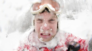 Einschneiter junger Mann mit Schneebrille und Schnee auf der Kleidung: Das Gesicht ist abgehärtet, weil weite Teile des Gesichts kaum behaart sind. Das Gesicht ist daher nicht so kälteempfindlich wie der Rest des Körpers und wir bekommen dort auch keine Gänsehaut. Das Gesicht is die letzte Stelle des Körpers, die wir im Winter verhüllen. 