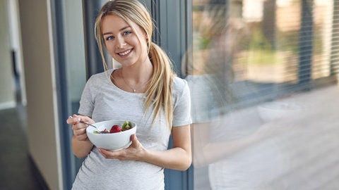 Junge schwangere Frau isst Obst aus einer Schale: Zu einer guten Ernährung gehören viel Gemüse, Obst, Vollkornprodukte, fettarme Milche und fettarme Milchprodukte. Milch und fettarmes Fleisch sind wichtig in der Schwangerschaft. Außerdem empfiehlt sich der Verzehr von Meeresfisch. 