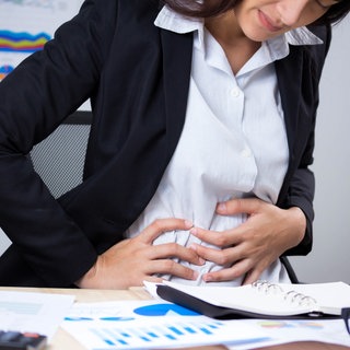 Frau in Business-Outfit sitzt am Schreibtisch und hält sich mit schmerzverzerrtem Gesicht den Bauch. Was sind Ursachen für starke Blähungen? Welche Hausmittel helfen?