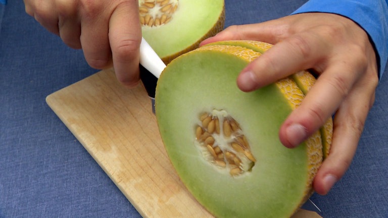 Hand mit Messer schneidet innen grüne, außen blassgelbe Galiamelone auf. Alles Wissenswerte zu Honigmelonen, Zuckermelonen, Wassermelonen, Galia-, Netzmelone und Cantalup
