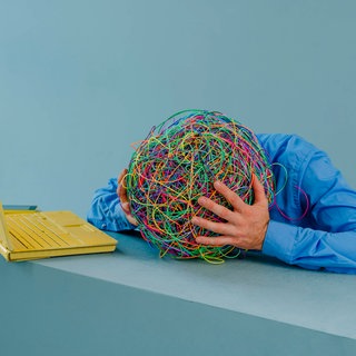 Symbolbild für ADHS wie es in der Wissenschaft und auf Social Media diskutiert wird: Person sitzt vor einem Laptop und hält den Kopf in den Händen, wobei der Kopf durch ein buntes Knäuel aus Gummibändern dargestellt wird