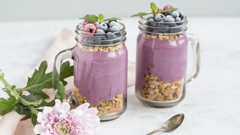 Zwei Gläser mit Joghurt, der u.a. gefrorene Aroniabeeren und anderes Obst enthält