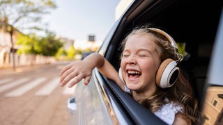 Mädchen mit Kopfhörer schaut aus Autofenster. Fröhliche Musik könnte helfen, sich von Reisekrankheit zu erholen. Und das Abspielen trauriger Musik kann Reiseübelkeit sogar noch triggern.