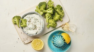 Brokkoliröschen auf einem Holzbrett, daneben Quarktip in Schälchen und Zitronensaft-Presse. Rezept für Brokkoli-Rohkost mit Dip.