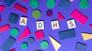 Das Wort "ADHS" auf Holzplättchen umbringt von bunten Formen. Kinder und Erwachsene können von ADHS betroffen sein.