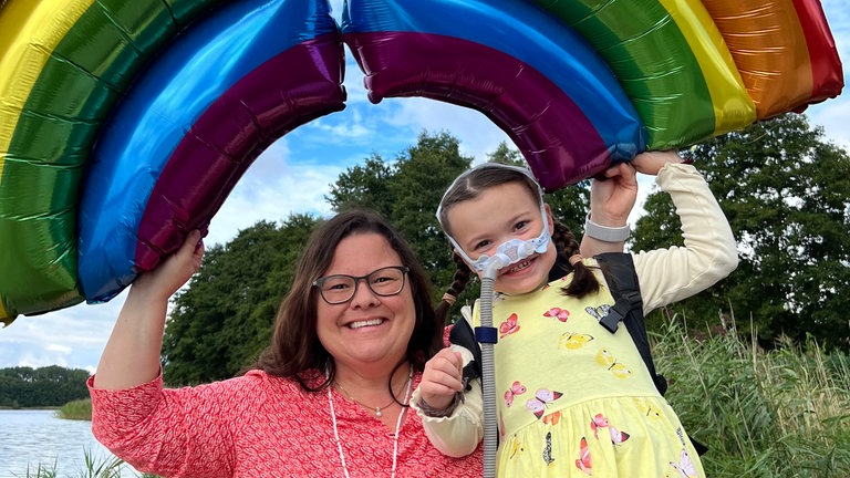 SWR1 Neuanfang: Lenya und ihre Mutter Sabine halten am Ufer eines Sees einen aufblasbaren Regenbogen in die Luft. Regenbogen hat Lenya Zeit ihres Lebens geliebt. An ihrem Todestag wurde einer sogar zum Symbol.