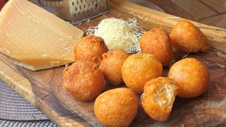 Goldgelbe Käsebällchen liegen auf einem Holzbrett, zusammen mit geriebenem Käse und einer Käsereibe. SWR1 Pfännle Rezept für goldgelbe Käsebällchen mit nur zwei Zutaten: Das ist ein schneller, knuspriger und unkomplizierter Snack oder eine leckere Beilage.